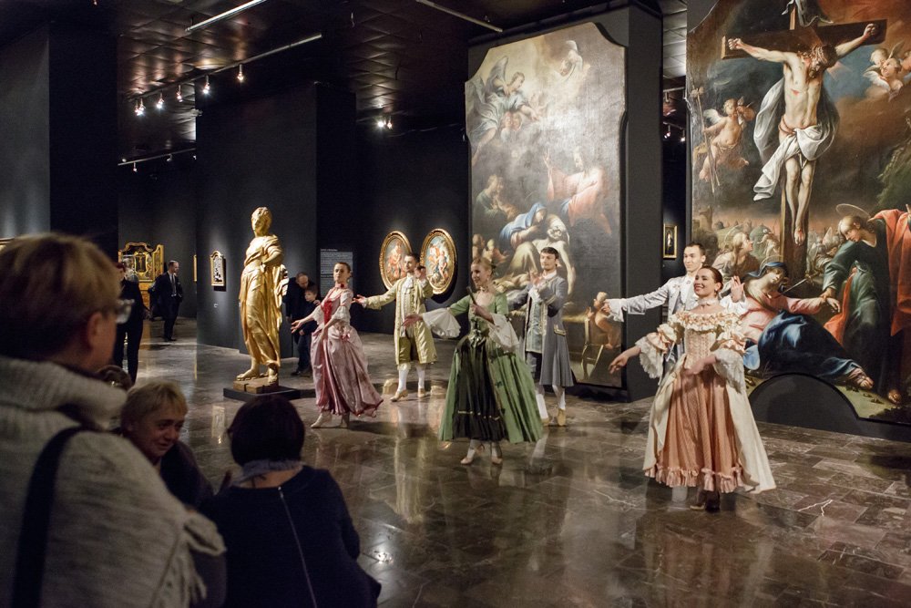 Grupa osób w historycznych kostiumach występuje w sali galerii przed wielkoformatowymi obrazami religijnymi i złotą rzeźbą, a w pierwszym planie widoczni są oglądający.