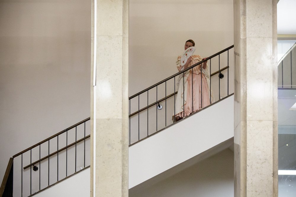 Osoba w historycznym różowo‑kremowym, długim stroju i pelerynie stoi na wewnętrznych schodach za metalową balustradą, przytrzymując ozdobny wachlarz przed twarzą; po bokach widoczne jasne marmurowe filary i gładkie beżowe ściany.
