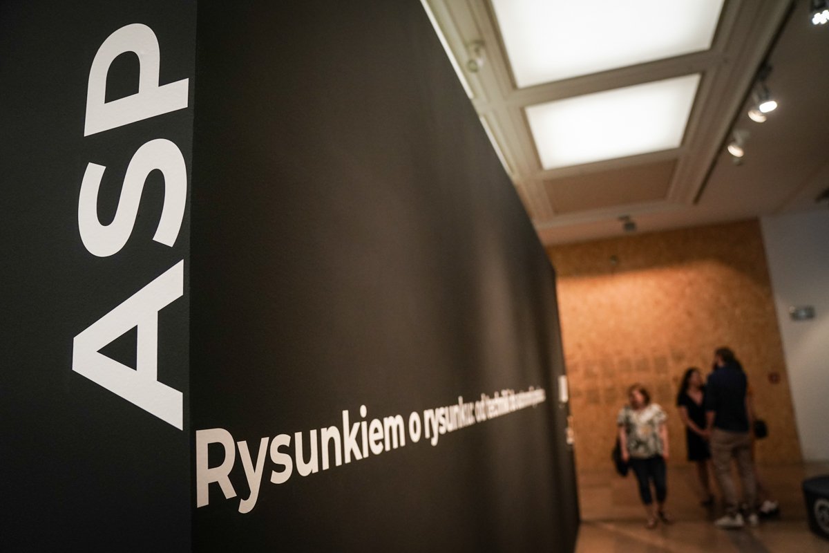 Wnętrze galerii z czarną ścianą wystawową na pierwszym planie z dużymi białymi pionowymi literami "ASP" i poziomym napisem "Rysunkiem...", w tle podświetlony sufit, drewniana ściana i trzech zamazanych zwiedzających.