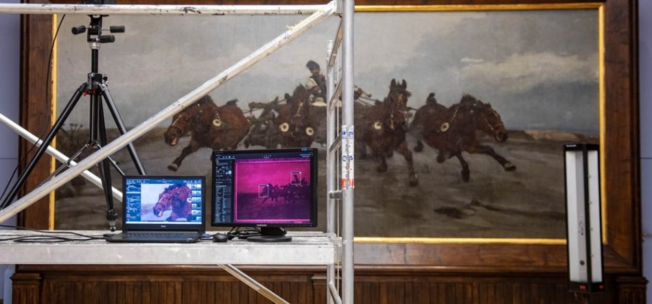 Metalowe rusztowanie ustawione przed dużym, złoconym, panoramicznym obrazem przedstawiającym galopujące konie i jeźdźców; na półce rusztowania stoją dwa monitory laptopa — lewy z niebieskim widokiem konia, prawy z różowo-fioletowym obrazem — po lewej widać statyw z zamontowanym urządzeniem, po prawej pionowy panel świetlny, a u dołu widoczna jest ciemna drewniana boazeria.