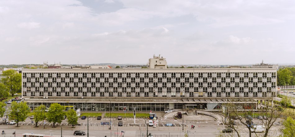 Panoramiczny, frontowy widok na duży prostokątny budynek o powtarzalnej, szachownicowej elewacji z wieloma kondygnacjami okien, przeszklonym parterem i zadaszonym wejściem; przed budynkiem parking i ulica z zaparkowanymi samochodami, niewielkie rondo, kilka osób oraz drzewa, a w tle rozległe, zachmurzone niebo.