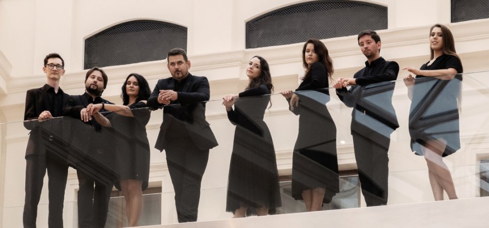 Ośmioro dorosłych w formalnych czarnych strojach stoi w linii za szklaną balustradą wewnętrznego balkonu, na jasnym tle z trzema łukowymi wnękami w ścianie.