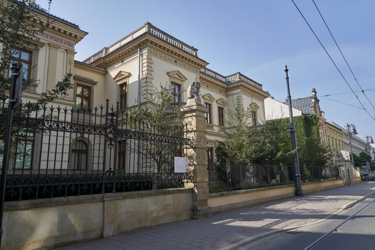 Widok z ulicy na kremowy neoklasycystyczny budynek z ozdobnymi gzymsami i balustradami, dużymi drewnianymi oknami, rzeźbą umieszczoną na kamiennym słupie przy ogrodzeniu, kutym żelaznym płotem na pierwszym planie, drzewami, latarnią uliczną i widocznymi nad ulicą liniami tramwajowymi pod jasnym, niebieskim niebem.