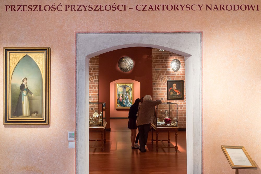Widok wnętrza muzeum z dużym napisem „PRZESZŁOŚĆ PRZYSZŁOŚCI – CZARTORYSCY NARODOWI” u góry, różowawą ścianą z kamiennym, łukowym przejściem prowadzącym do ceglastego wnętrza galerii, gdzie wiszą oprawione obrazy, stoją szklane gabloty na drewnianych stolikach, po lewej wizerunek kobiety w długiej sukni, po prawej tablica informacyjna na stojaku i dwóch zwiedzających, z których jedna osoba z siwymi włosami pochyla się nad gablotą.