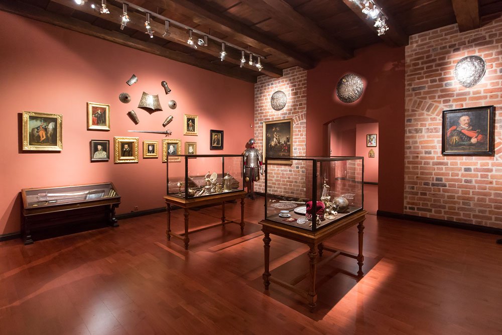 Galeria muzealna o bordowych i ceglanych ścianach, drewnianej podłodze i belkowanym suficie, oświetlona reflektorami, z dwiema oszklonymi witrynami na rzeźbionych drewnianych nogach w centrum, stojącą zbroją przy tylnej ścianie, licznymi obrazami w złotych ramach, wiszącymi elementami zbroi i dekoracyjnymi okrągłymi tarczami oraz łukowym przejściem do sąsiedniego wnętrza.
