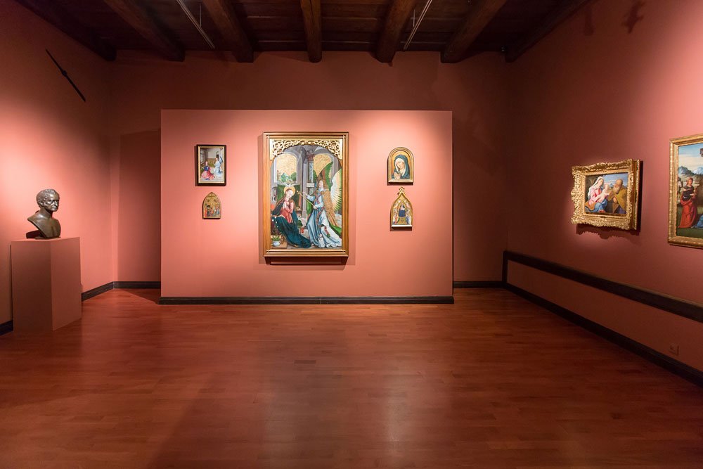 Wnętrze galerii z różowawymi ścianami i drewnianą podłogą, centralnie ustawiona ściana z pięcioma obrazami przedstawiającymi postacie (duży obraz w środku i cztery mniejsze wokół), po lewej na postumencie brązowe popiersie, po prawej dodatkowe obrazy w złoconych ramach oraz widoczne drewniane belki stropu i puste centralne przestrzenie podłogi.