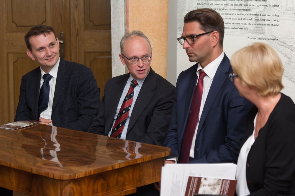 Cztery formalnie ubrane osoby siedzą przy błyszczącym drewnianym stole; od lewej trzech mężczyzn w garniturach i krawatach (dwóch w okularach), po prawej kobieta z krótkimi blond włosami trzymająca dokumenty, w tle drewniane drzwi i plansza z tekstem.