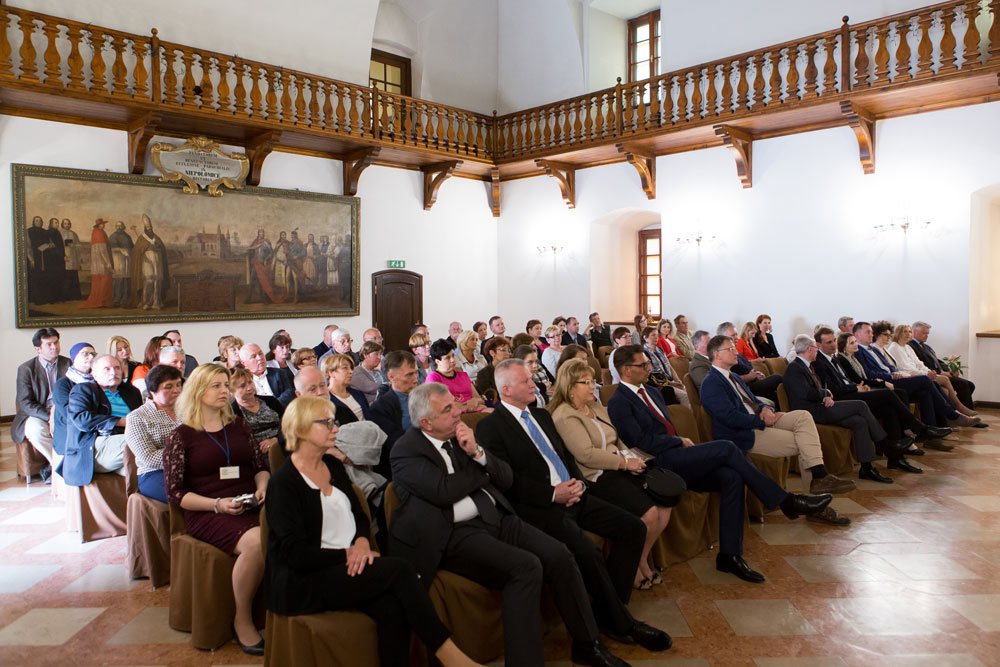 Wnętrze dużej, jasnej sali z ozdobną drewnianą galerią i dużym malowidłem na ścianie, wypełnione kilkudziesięcioma formalnie ubranymi osobami siedzącymi w kilku rzędach i zwróconymi w stronę prawej części kadru.