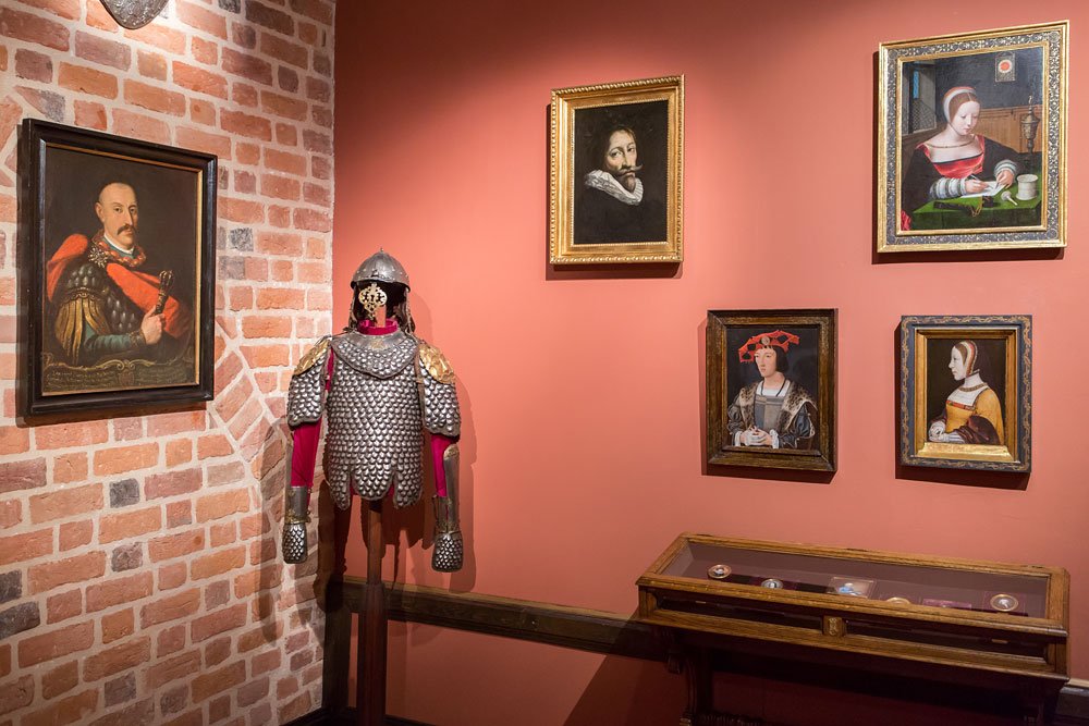 Galeria z ceglaną ścianą po lewej i łososiową po prawej, na których rozmieszczono kilka oprawionych portretów, obok stoi na stojaku metalowa zbroja łuskowa z hełmem i czerwonym materiałem pod spodem, a po prawej niska drewniana witryna ze szklanym blatem z drobnymi eksponatami.