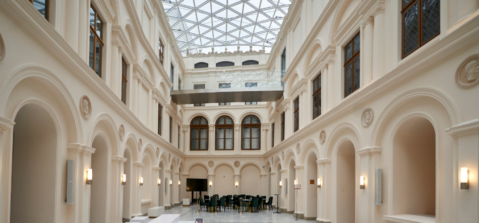 Przestronne, jasne atrium z dwiema kondygnacjami klasycznych arkad i pilastrów po obu stronach, dekoracyjnymi medalionami w ścianach, dużym przeszklonym dachem kratownicowym, wiszącym metalowym przejściem oraz stolikami i krzesłami ustawionymi na jasnej marmurowej posadzce.