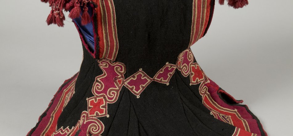 Fragment czarnego, rozkloszowanego stroju ozdobionego czerwonymi pasami i bordowymi, geometryczno‑falistymi aplikacjami obramowanymi złotą nicią i małymi złotymi ćwiekami, z widoczną niebieską podszewką i czerwonymi frędzlami u góry.