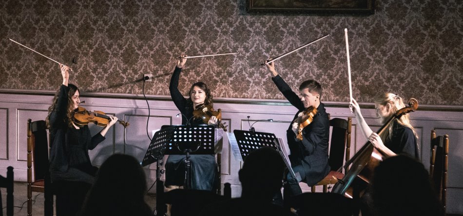 Na niewielkiej scenie w sali z ozdobną tapetą i boazerią cztery siedzące osoby w ciemnych ubraniach trzymają instrumenty smyczkowe — trzy mniejsze (skrzypce/alt) i jedną wiolonczelę — stoją przed pulpitami z nutami i jednocześnie unoszą smyczki nad głowami, a z przodu kadru widać ciemne sylwetki publiczności.