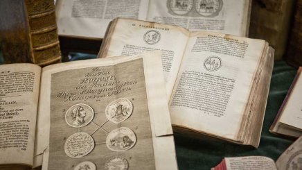 Kilka otwartych starych książek rozłożonych na zielonym materiale; na pierwszym planie widoczna strona z ozdobnym tytułem i sześcioma okrągłymi miedzorytowymi wizerunkami, w tle kolejne otwarte tomy i stojące grzbiety.
