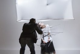 Fotograf w ciemnej odzieży z aparatem schylający się przed osobą w jasnej bluzce i ciemnej spódnicy stojącą przy instalacji z metalowych prętów pod dużym prostokątnym softboxem w jasnym studiu na szarym tle.