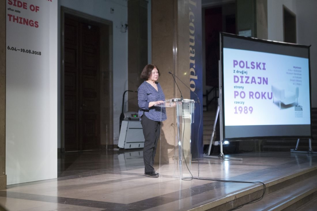 Kobieta stojąca przy przezroczystej mównicy z mikrofonem na podwyższeniu w dużej sali wystawowej, po prawej ekran projekcyjny z widocznym napisem "POLSKI DIZAJN PO ROKU 1989", po lewej wysoka biała plansza z datami "6.04–19.08.2018", marmurowa podłoga, kolumny i widoczne kable.