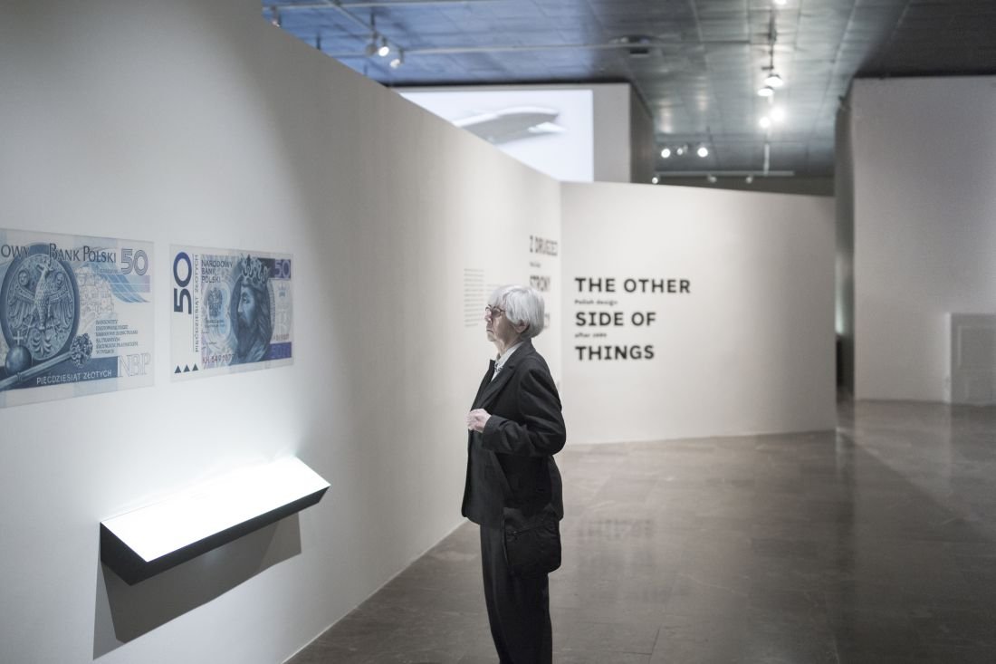 Osoba o białych włosach i ciemnym płaszczu stoi w jasnej galerii przed ścianą z powiększonymi wizerunkami banknotów 50 zł i podświetloną gablotą; w tle duży napis THE OTHER SIDE OF THINGS i przestronne, oświetlone wnętrze muzeum.