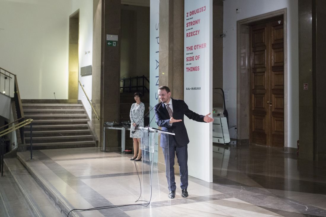 Na marmurowej posadzce dużego hallu mężczyzna w garniturze stoi za przezroczystą mównicą z mikrofonem, gestykuluje wyciągniętą dłonią; za nim po lewej widać schody, po prawej wysokie drewniane drzwi i panel wystawowy z czerwonym napisem Z DRUGIEJ STRONY RZECZY / THE OTHER SIDE OF THINGS, a przy ścianie stoi kobieta w wzorzystej sukience obok sprzętu i leżących kabli.