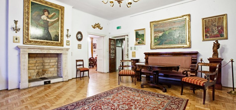 Wnętrze stylowego salonu z parkietem i ornamentowym dywanem na pierwszym planie, po lewej kamienny kominek z dużym złoconym obrazem nad nim przedstawiającym postać kobiecą, po prawej drewniana sofa i krzesła w pasiastym obiciu z niskim stolikiem, na ścianach kilka oprawionych obrazów, rzeźba na cokole, żyrandol sufitowy i otwarte drzwi prowadzące do sąsiedniego pomieszczenia.