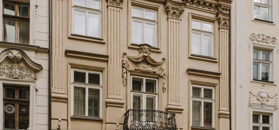 Fragment elewacji zabytkowej kamienicy w odcieniach beżu i kremu z pionowymi pilastrami i rzeźbionymi detalami, centralnym prostokątnym oknem z czarnym, ażurowym balkonem, nad nim dekoracyjnym kartuszem z motywem twarzy i draperii, oraz kilkoma białymi ramami okien i fragmentami sąsiednich zdobionych fasad po bokach.