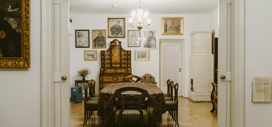 Widok przez otwarte podwójne drzwi na jadalnię z centralnym stołem przykrytym wzorzystym obrusem i kilkoma drewnianymi krzesłami, nad stołem żyrandol; na tylnej ścianie ozdobna intarsjowana komoda otoczona oprawionymi rysunkami i portretami, po prawej biała kaflowa piec, po lewej fragment dużego złoconego obrazu i mały stolik z suchym bukietem, jasna parkietowa podłoga.