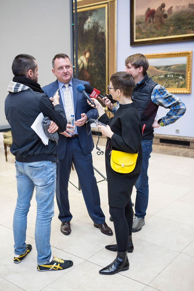 Mężczyzna w granatowym garniturze i jasnym krawacie, trzymający butelkę wody, otoczony przez trzech dziennikarzy trzymających mikrofony i notatki w sali z obrazami wiszącymi na ścianach.