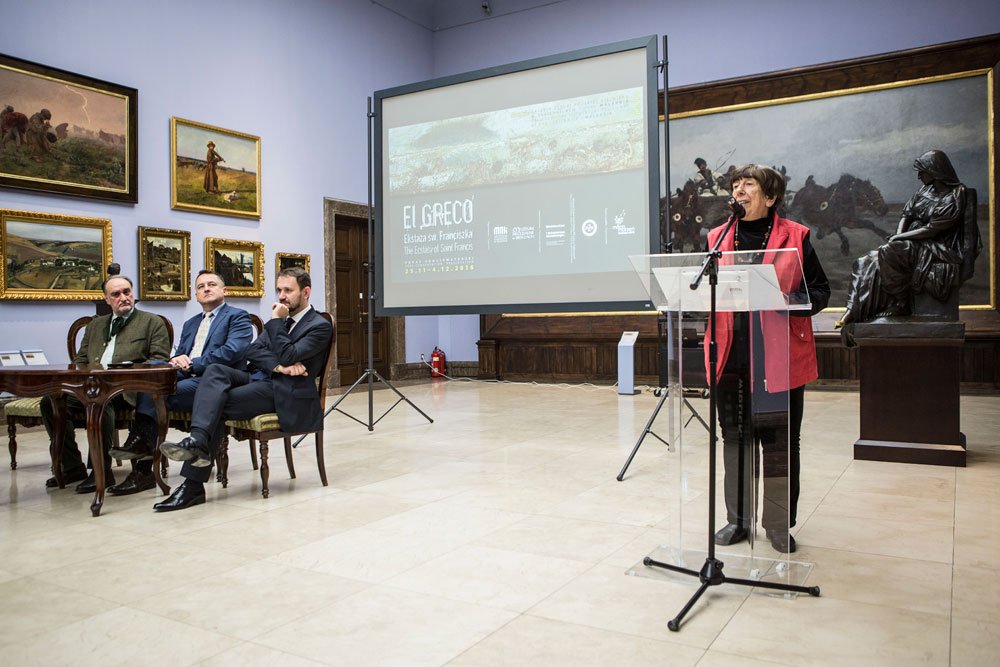 Kobieta w czerwonej kamizelce stoi przy przezroczystej mównicy z mikrofonem po prawej stronie zdjęcia, za nią ekran z projekcją z napisem "EL GRECO", po lewej trzy siedzące osoby przy drewnianym stoliku, a w tle ściany z wieloma oprawionymi obrazami i duża ciemna rzeźba na cokole.