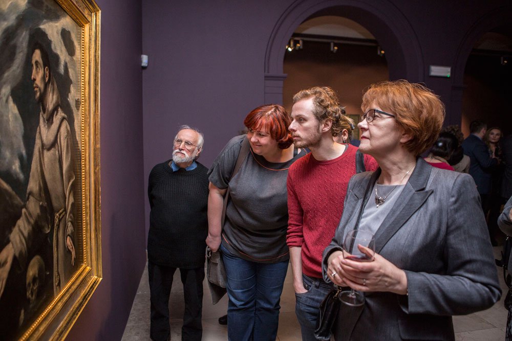 Grupa czterech osób stoi przy fioletowej ścianie galerii, wpatrując się w duży obraz w złotej ramie po lewej; z przodu kobieta w szarej marynarce trzyma kieliszek wina, obok stoi mężczyzna w czerwonym swetrze, pochyla się kobieta z krótkimi rudymi włosami w szarej bluzce, a za nimi stoi starszy siwy mężczyzna w czarnym swetrze, w tle widać łuk wejściowy i innych zwiedzających.