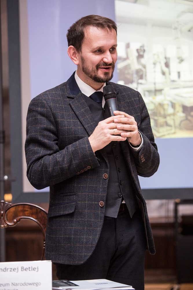 Mężczyzna w marynarce w kratę, kamizelce, białej koszuli i krawacie stoi przed ekranem projekcyjnym i trzyma oburącz mikrofon, obok widać drewniane krzesło oraz stół z dokumentami i tabliczką z nazwiskiem.