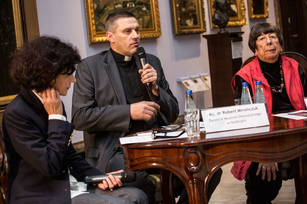Trzy osoby siedzą przy ozdobnym drewnianym stoliku podczas spotkania: mężczyzna w szarym garniturze z koloratką mówi przez mikrofon, po lewej kobieta trzyma drugi mikrofon, po prawej starsza kobieta w czerwonej kamizelce, na stole butelki wody, szklanka i kartka z nazwiskiem, w tle obrazy w ramach i popiersie.