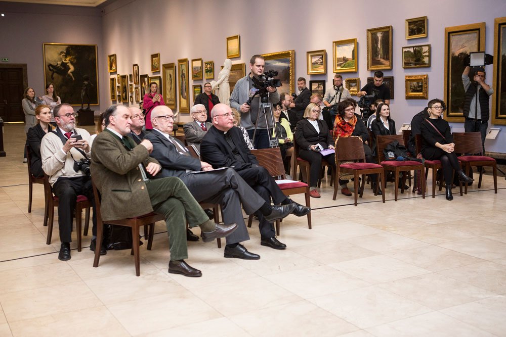 Kilkanaście osób siedzi na drewnianych krzesłach z czerwonymi siedziskami w przestronnej sali galerii z jasną posadzką i fioletowymi ścianami ozdobionymi wieloma oprawionymi obrazami i stojącą rzeźbą, a z tyłu stoją kamerzyści i fotografowie z kamerami i aparatami.