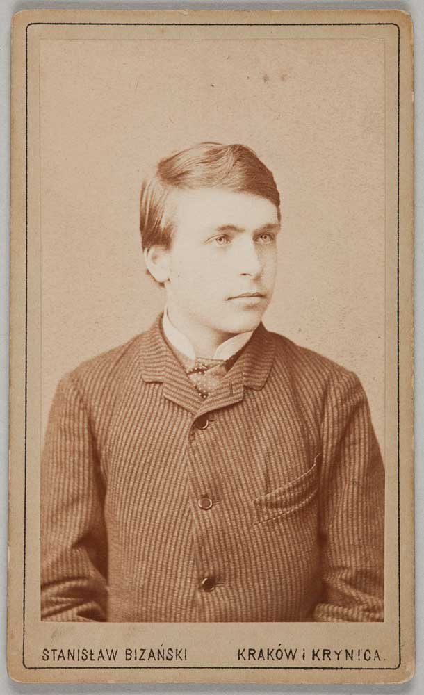 Sepia portret studyjny na karcie fotograficznej przedstawiający młodego mężczyznę od klatki piersiowej w górę, zwróconego nieco w prawo, w prążkowanym żakiecie i apaszce, z krótkimi zaczesanymi włosami i neutralnym wyrazem twarzy, z cienką ramką i drukowanym napisem na spodzie: "STANISŁAW BIZAŃSKI KRAKÓW i KRYNICA".