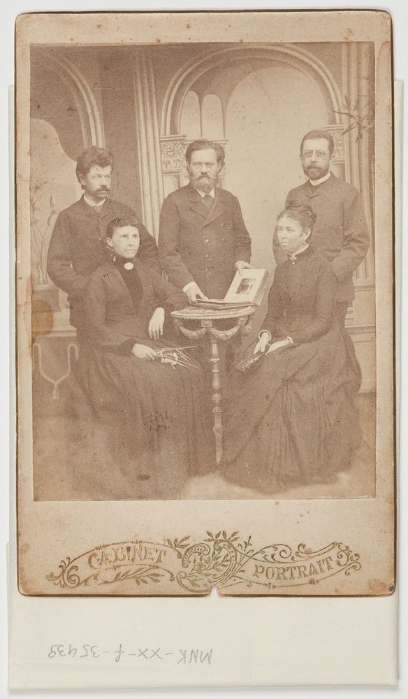 Sepiowe cabinet card przedstawiające pięć osób: trzy stojące osoby męskie w tle i dwie siedzące kobiety z przodu przy małym okrągłym stoliku z otwartym albumem, na ozdobnym malowanym tle z arkadami, z ornamentową dolną ramką z napisem "Cabinet Portrait" oraz widocznymi przebarwieniami i śladami zużycia.