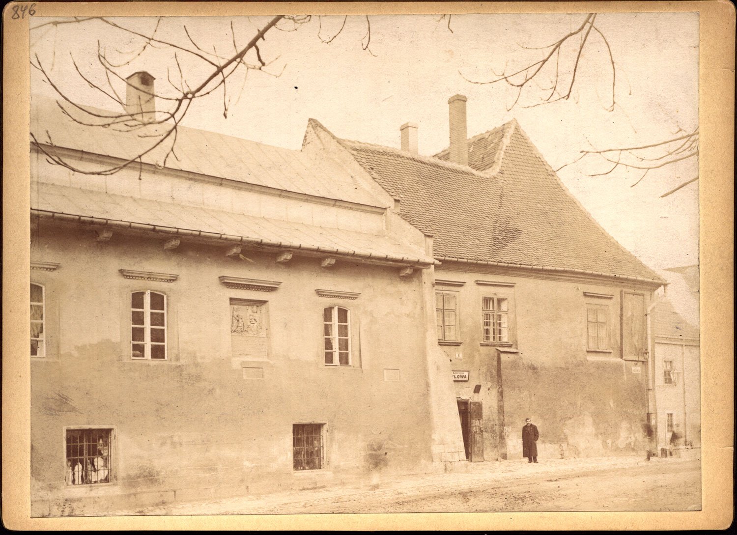 Fotografia w sepii przedstawiająca fasadę starego budynku z dużym, spadzistym dachem krytym dachówką i niższą, metalową częścią dachu po lewej, szeregiem prostokątnych okien (w tym kraty w przyziemiu), dwoma kominami, gałęziami drzewa nachodzącymi z góry kadru oraz jedną osobą w ciemnym płaszczu stojącą przy wejściu pod małym szyldem, przed budynkiem widoczna ulica.