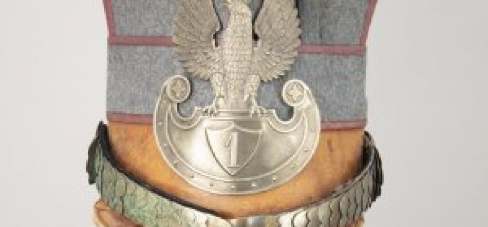 Fragment szarej filcowej czapki z czerwonym obszyciem, brązowym skórzanym daszkiem i srebrną metalową plakietą przedstawiającą orła z rozpostartymi skrzydłami nad półokrągłą tarczą z cyfrą "1", u dołu ozdobiony metalowym paskiem i zielonym wieńcem.