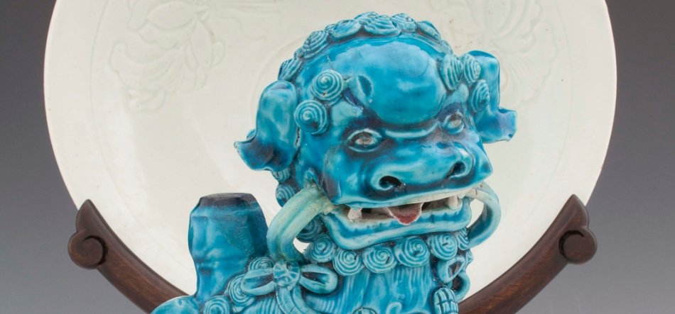 Turkusowa szkliwiona ceramiczna figurka lwa strażniczego z spiralną grzywą, otwartym pyszczkiem z widocznymi zębami i kółkiem w pysku, ustawiona przed dużym białym talerzem z wytłoczonym wzorem na ciemnobrązowym drewnianym stojaku.