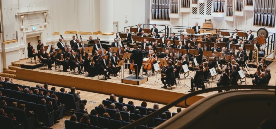 Orkiestra symfoniczna w ciemnych strojach zajmuje drewnianą scenę sali koncertowej z dyrygentem stojącym na podwyższeniu, sekcją smyczków z przodu, dętymi blaszanymi i perkusją z tyłu, dużymi piszczałkami organów na tylnej ścianie oraz widownią siedzącą w fotelach i balkonem na pierwszym planie.