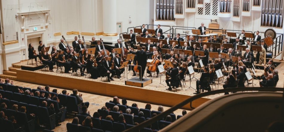 Duża orkiestra na drewnianej scenie sali koncertowej o białych ścianach z złotymi detalami: dyrygent stoi na podium, wokół siedzą muzycy z sekcjami smyczkową, dętą i perkusyjną, za nimi widoczne piszczałki organów, a na pierwszym planie siedząca publiczność w rzędach foteli.