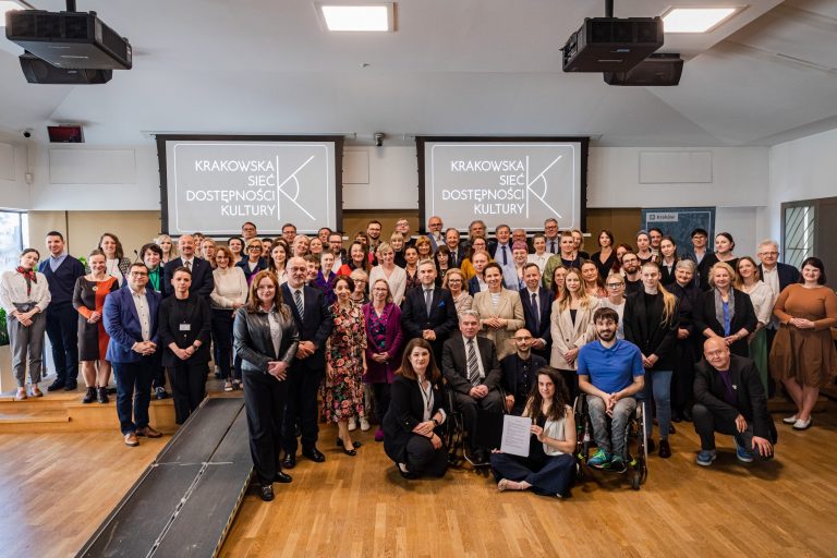 Duże zdjęcie grupowe kilkudziesięciu osób ustawionych w kilku rzędach na scenie i przed nią w sali konferencyjnej z dwiema dużymi projekcjami na ścianie z napisem „KRAKOWSKA SIEĆ DOSTĘPNOŚCI KULTURY” i logo, sufit z projektorami, drewniana podłoga, z przodu kilka osób siedzi, jedna kobieta trzyma dokument, u dołu po prawej widoczna osoba na wózku inwalidzkim.