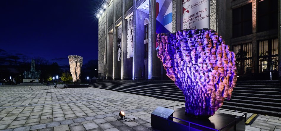 Plac z kwadratową posadzką przed monumentalnym budynkiem z kolumnami i dużymi banerami wieczorem, na pierwszym planie fioletowo-pomarańczowo oświetlona chropowata rzeźba na cokole, dalej druga oświetlona rzeźba, schody prowadzące do wejścia, reflektory i ciemne niebo w tle.