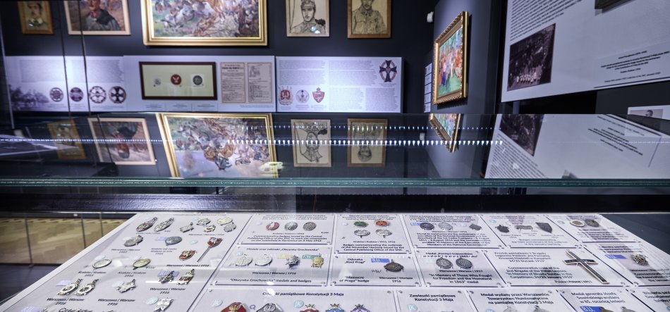 Wnętrze muzeum z przeszkloną gablotą na pierwszym planie, w której na planszach leżą starannie ułożone medale, odznaki i karty opisowe, a w tle na ciemnej ścianie powieszone ramowane obrazy, portrety i teksty informacyjne oraz ich odbicia na szklanej tafli.