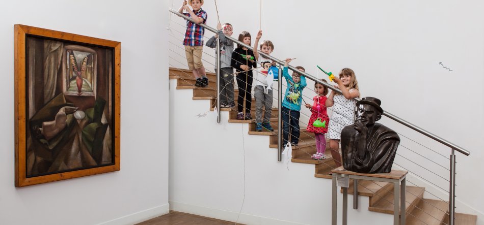 Wnętrze galerii z białymi ścianami i drewnianą podłogą; po lewej duży oprawiony obraz na ścianie, po prawej na stoliku ciemne popiersie w kapeluszu, a na drewnianych schodach z metalową poręczą stoi kilka dzieci w rzędzie trzymających zabawki, papierowe chorągiewki i pistolety na wodę.