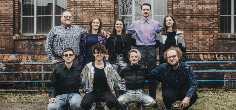 Grupowe zdjęcie dziewięciu osób ustawionych w dwóch rzędach przed ceglanym budynkiem z dużymi prostokątnymi oknami; w tyle pięć osób stoi z rękami na ramionach sąsiadów, z przodu cztery osoby kucają obok niebieskiej ławki na trawie, wszyscy w różnorodnych strojach (m.in. błyszczące srebrne spodnie, czarna puchowa kurtka, wzorzyste koszule, okulary).
