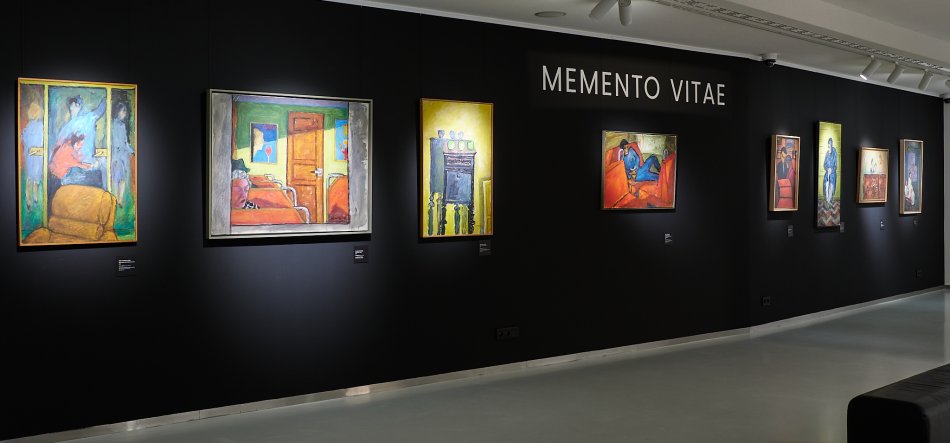 Wnętrze galerii z czarną ścianą wystawową, na której w rzędzie wiszą podświetlone kolorowe obrazy w ramach, nad środkową częścią ściany duży biały napis "MEMENTO VITAE", jasna podłoga, sufitowe reflektory i po prawej widoczne siedzisko.