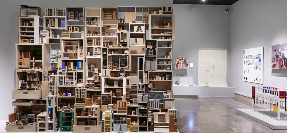 Wnętrze galerii z dużą, zajmującą lewą część kadru instalacją z wielu drewnianych, otwartych pudełek i miniaturowych modeli wnętrz, po prawej białe ściany z kilkoma obrazami, kolorową kurtką na ekspozytorze i dodatkowymi białymi podestami z rzeźbami, błyszcząca kamienna posadzka i ciemny sufit z reflektorami.