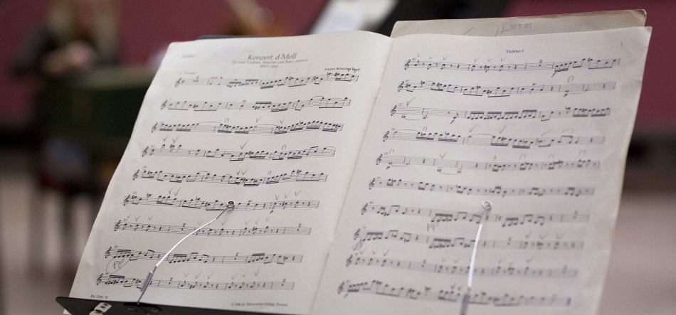 Otwarta partytura ustawiona na pulpicie nutowym, dwie strony z zapisanymi pięcioliniami i nutami (na lewej stronie widoczny tytuł "Konsert d Moll"), z ołówkowymi zapiskami i metalowymi klipsami trzymającymi kartki, na rozmytym tle widoczne niewyraźne postaci i meble.