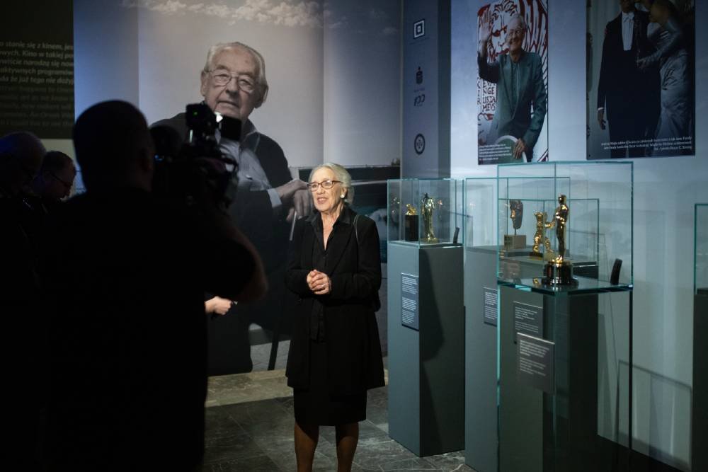 Kobieta w ciemnym ubraniu z dłońmi złożonymi stoi w sali wystawowej przed szklanymi gablotami z złotymi statuetkami, po lewej widoczna sylwetka operatora kamery, a w tle duże czarno-białe zdjęcie mężczyzny i plakaty na ścianie.