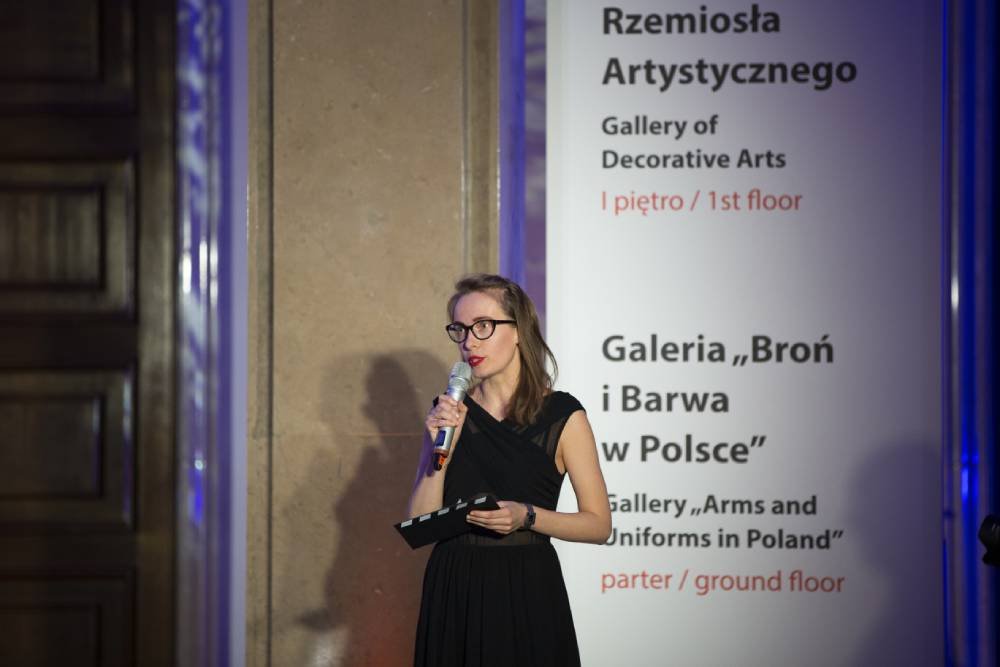 Kobieta w czarnej sukience i okularach, z czerwonymi ustami, trzyma mikrofon i notes stojąc przy filarze obok dużej planszy z napisami po polsku i angielsku o Galerii Rzemiosła Artystycznego i galerii „Broń i Barwa w Polsce”, po lewej widoczne drewniane drzwi i cień na filarze.