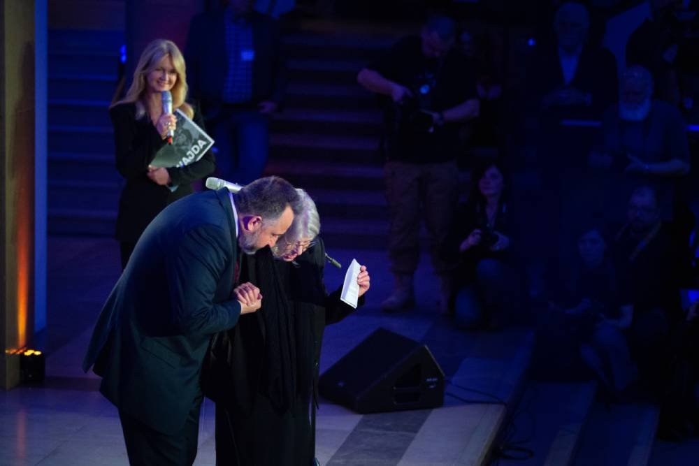 Na scenie mężczyzna w garniturze i starsza kobieta pochylają się trzymając się za ręce, kobieta ma w dłoni kartkę, obok stoi mikrofon, w tle blond kobieta z mikrofonem i teczką oraz ciemna publiczność i fotografowie oświetleni niebieskim światłem.