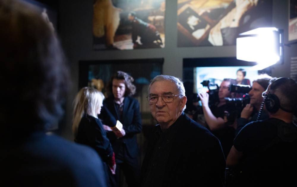 Starszy mężczyzna w okularach stoi na pierwszym planie w ciemnym pomieszczeniu, za nim po prawej kamerzyści z dużym jasnym oświetleniem, po lewej dwie osoby rozmawiające, a w tle powieszone na ścianie zdjęcia lub obrazy.