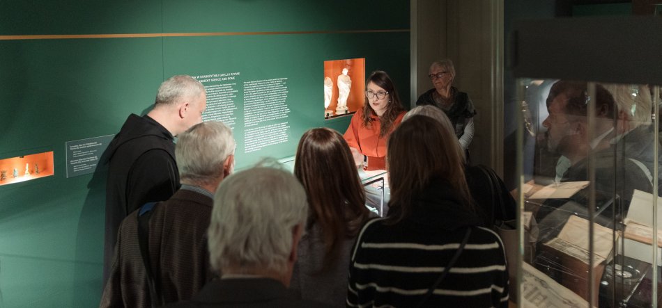 Wnętrze muzeum z zieloną ścianą z tekstami informacyjnymi i małymi podświetlonymi niszami, kobieta w czerwonej bluzce stoi przy przeszklonej gablocie, wokół niej grupa osób stojących tyłem ogląda eksponaty, po prawej widoczna kolejna przeszklona witryna z otwartymi książkami i odbiciem twarzy mężczyzny w szybie.